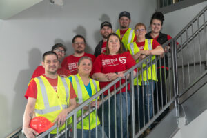 Team members at Dynamic Lifecycle Innovations wearing safety vests pose on the stairs.