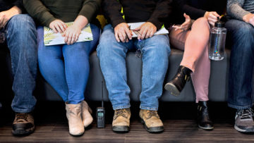 Photo of five employees sitting on a couch holding different items for their jobs - one holds a yellow hard hat and safety glasses, one holds a planner, another holds glasses, a cell phone, and notebook, another holds a water bottle, and the last person is holding safety glasses.