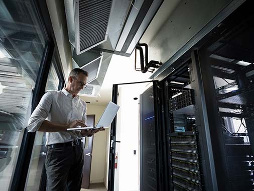 An IT engineer working on a laptop while standing in front of a large server rack.